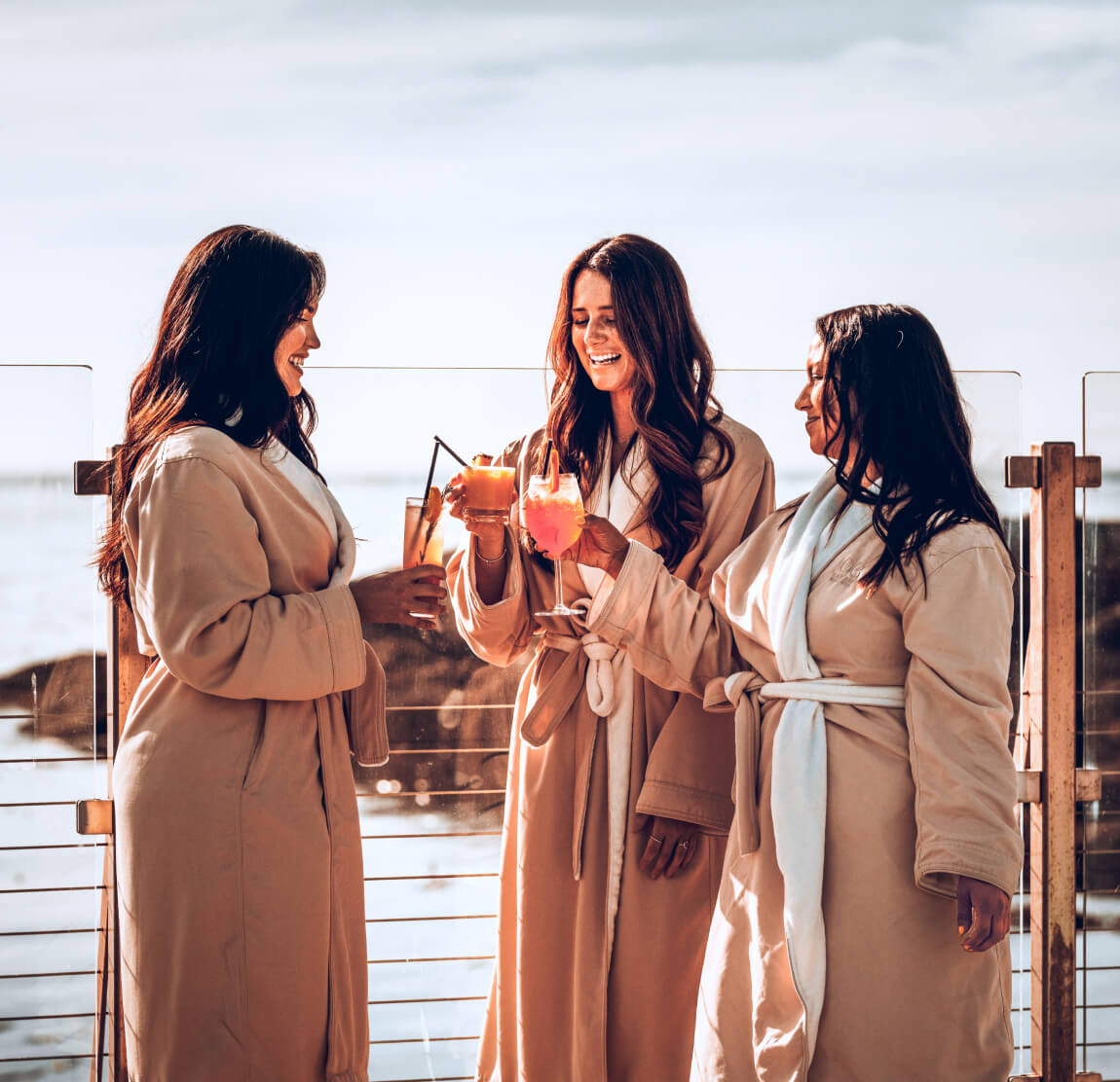 three young brunette women holding cocktails wearing robes pool side toasting ocean views