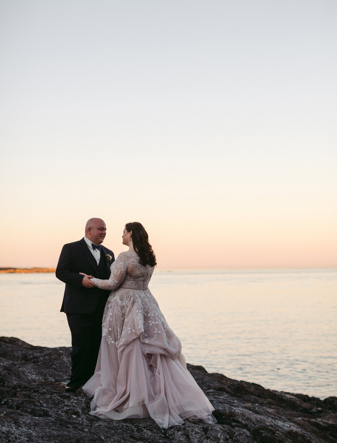 husband and wife gazing into each others eyes standing on oceanfront rock edge beautifully detailed wedding dress sunset