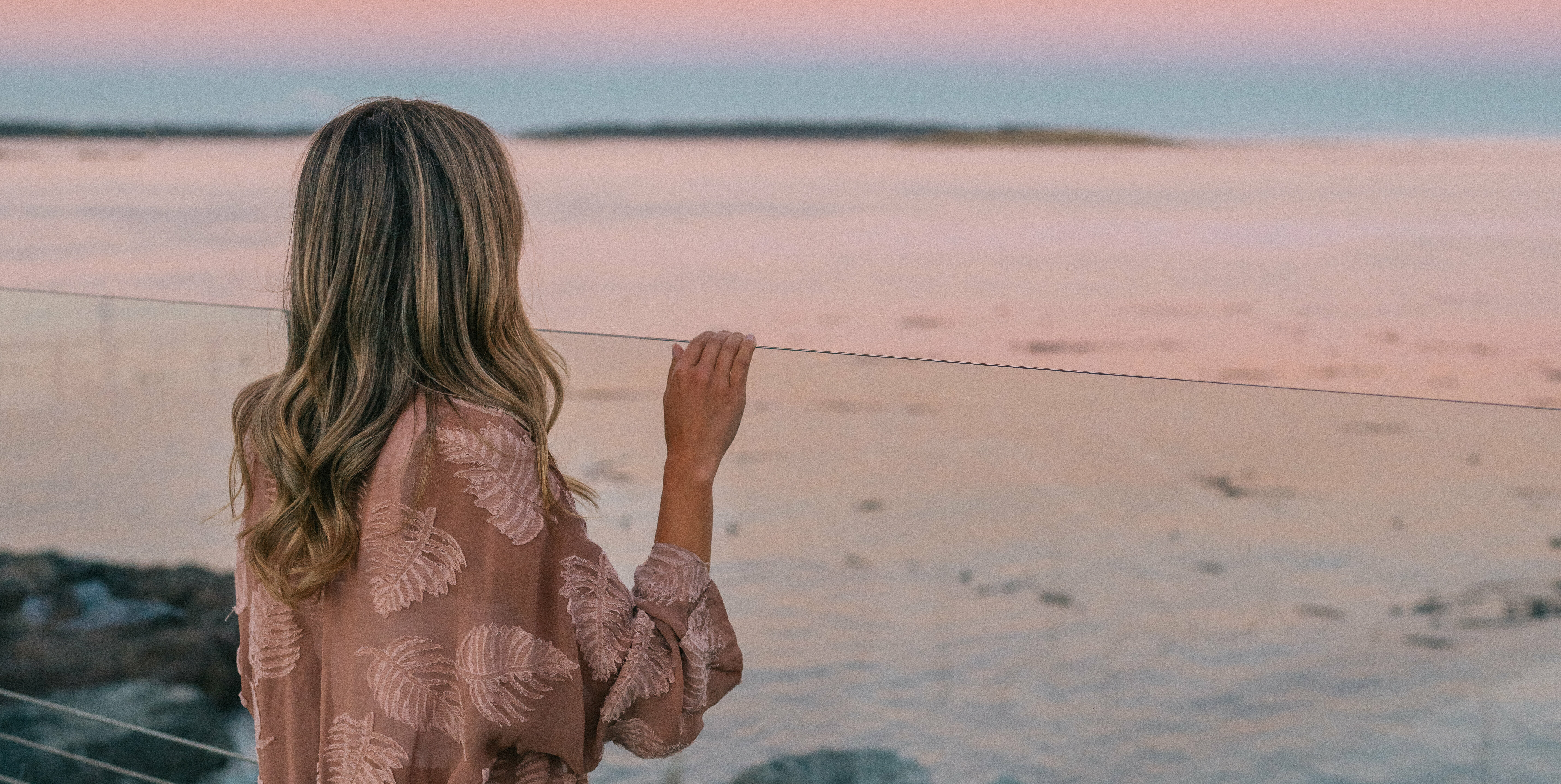 woman in pink jacket looking over glass railing to sunset on ocean